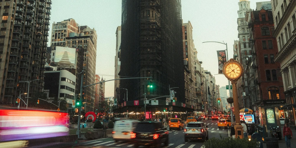 这是一条繁忙的城市街道，中心有被脚手架包裹的高楼，右侧有立柱钟表，街上车辆穿梭，光线显示为清晨或傍晚。